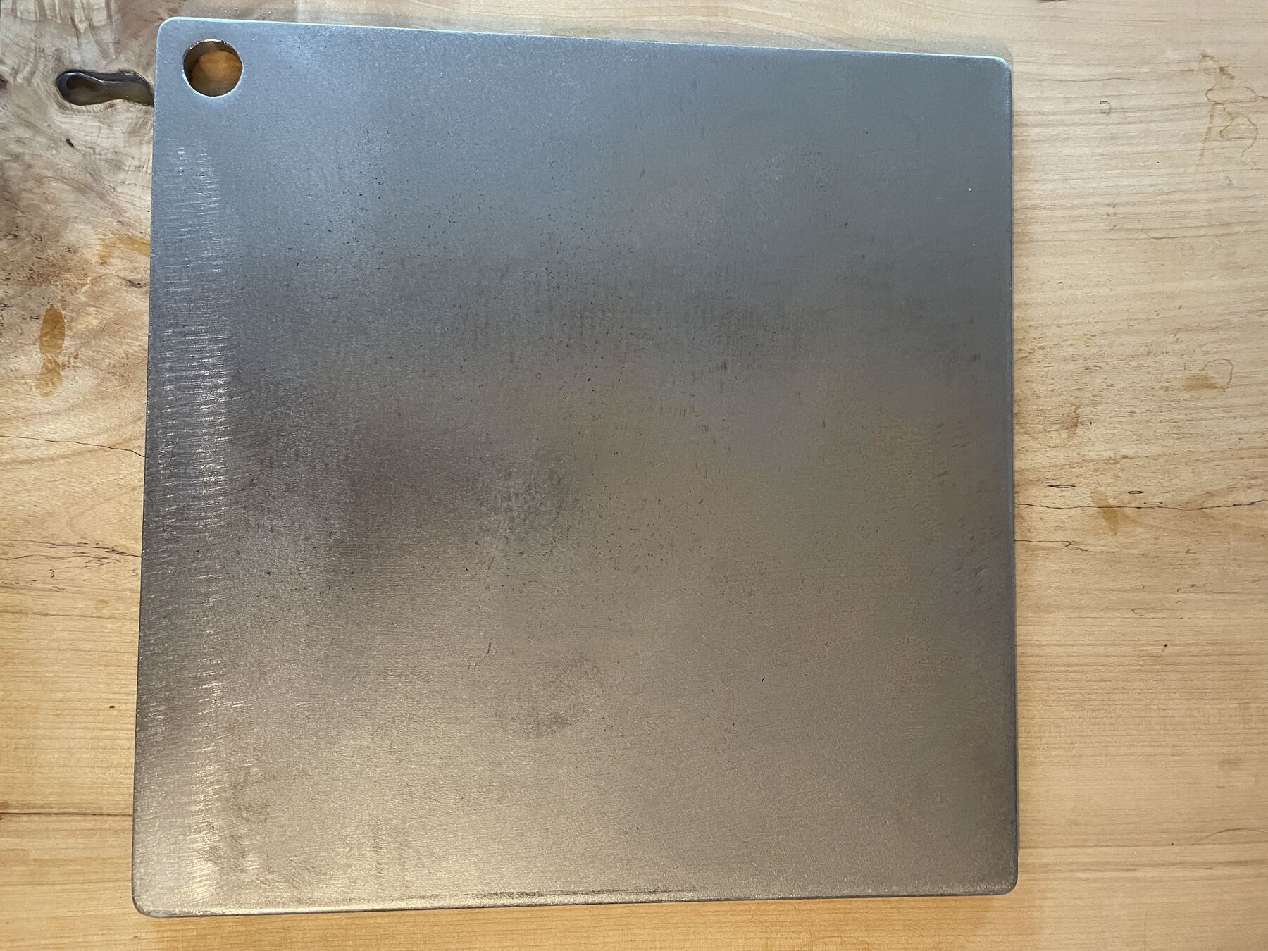 A 16 by 16 inch hand cut carbon steel pizza and baking steel on a maple slab.